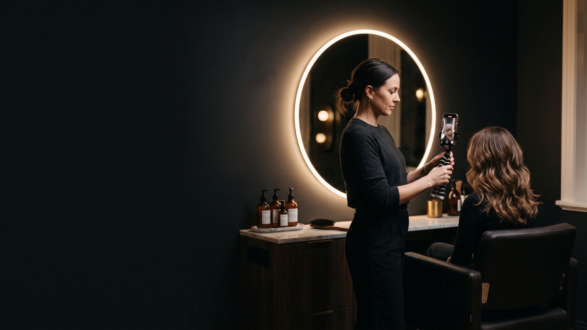 A stylist films a short video of her client's finished hair on a phone, in a dark salon lit by a halo mirror